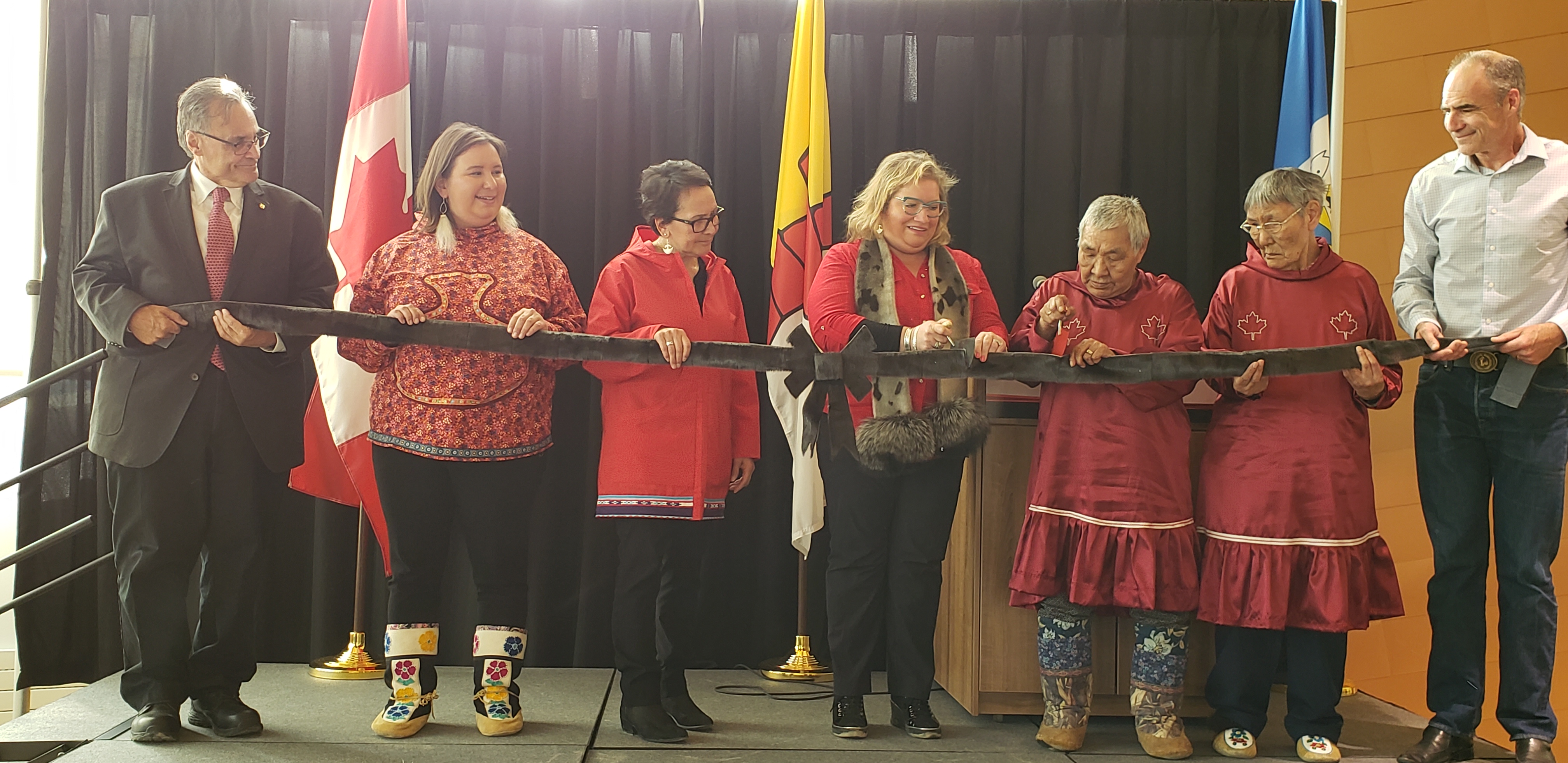 Photo – Three women holding a ribbon of seal skin. The woman on the right cuts it with an ulu. 